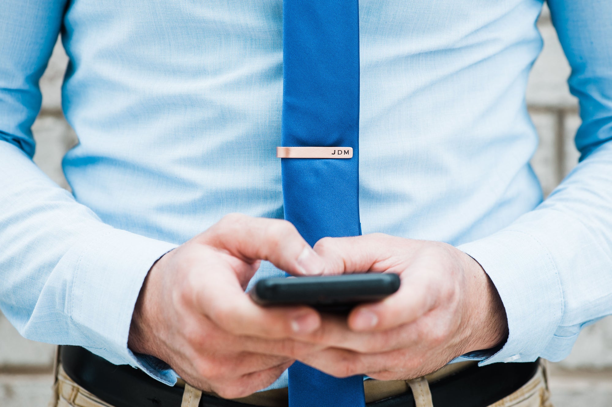 Man appears to be texting from his cell phone. He is wearing light blue shirt, dark blue tie. Attached to tie is a tie clip personalized with his initials.
